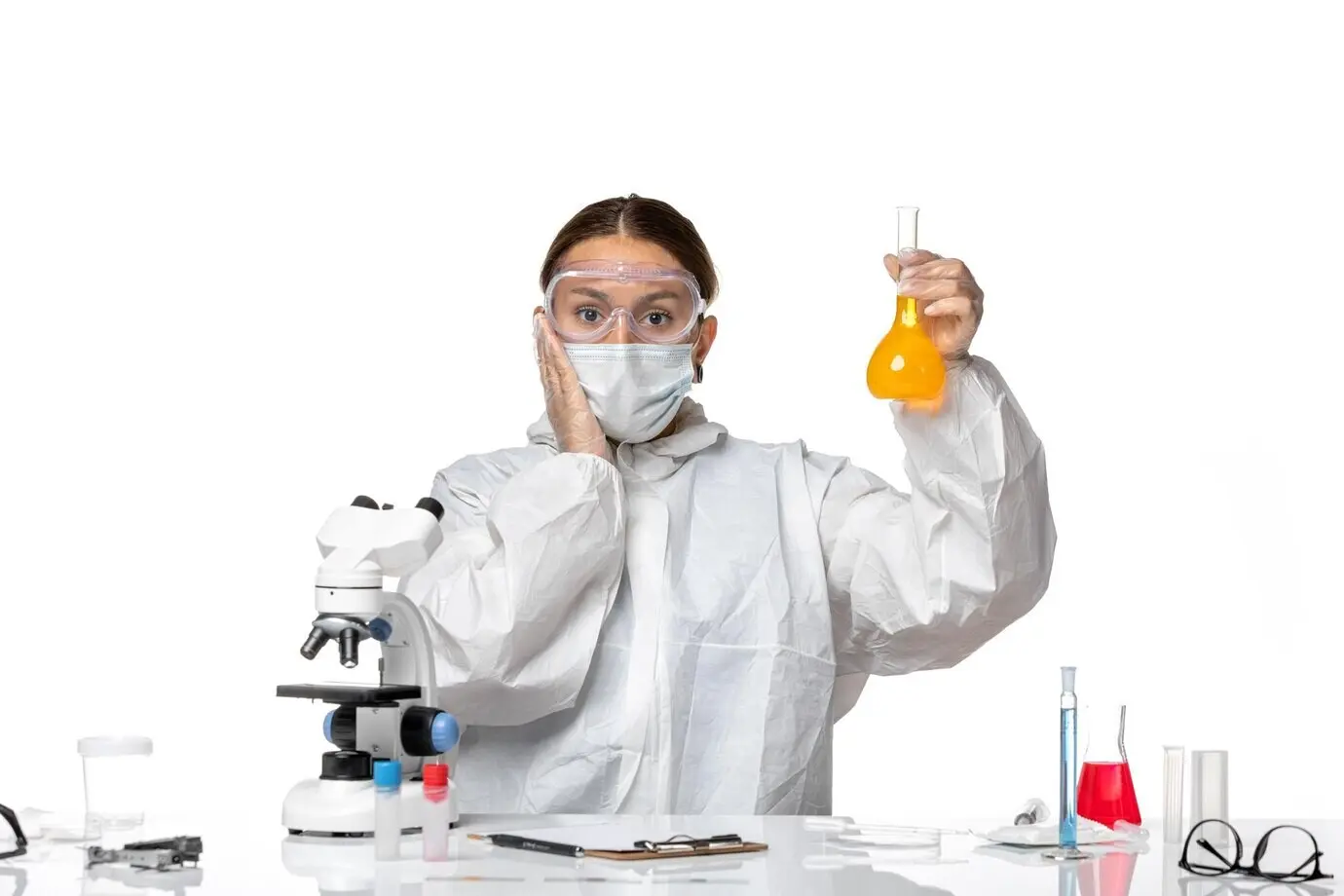 Vista frontal de una doctora con traje especial y mascarilla, sosteniendo un matraz con solución amarilla sobre un fondo blanco claro; covid, pandemia, salud, coronavirus, virus.